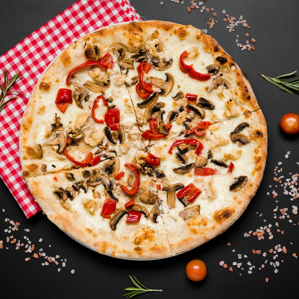 A white pizza with mushrooms, red peppers, and chicken, on a black surface with a red checkered napkin.
