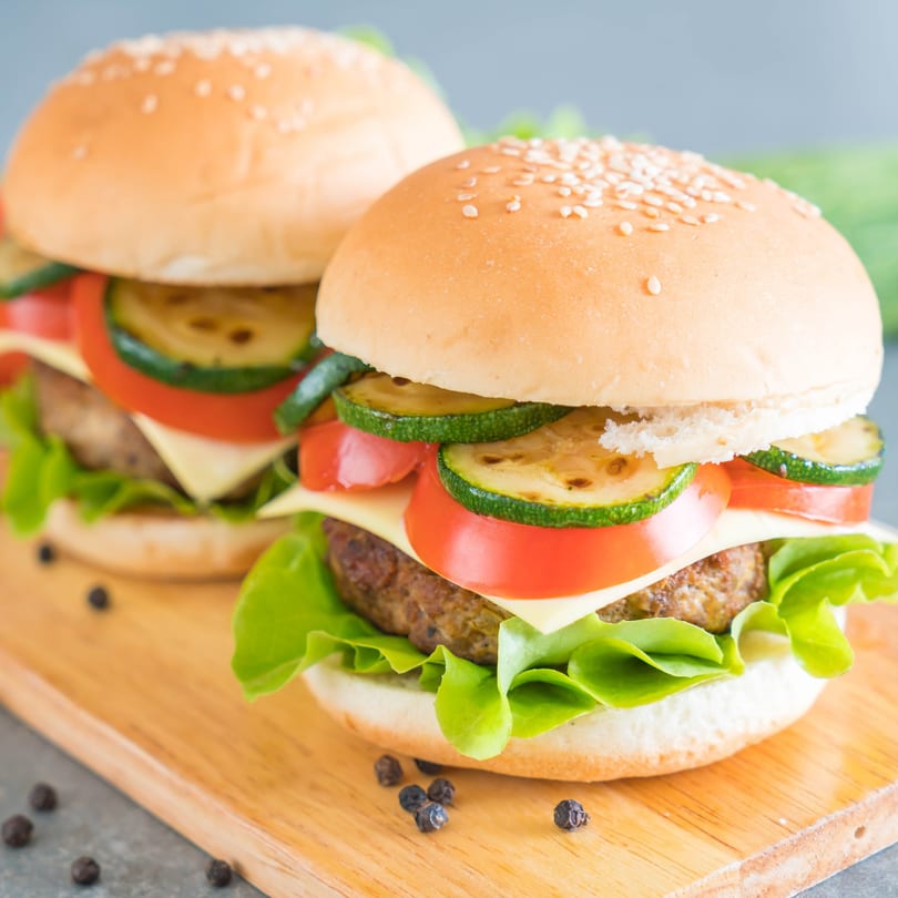 Two deli burgers with cheese, lettuce, tomato, and grilled zucchini on a wooden board.